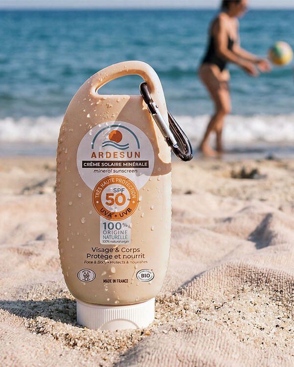 Flacon de crème solaire minérale SPF50 ARDESUN posé dans le sable sur une plage avec une personne jouant au ballon en arrière-plan flou